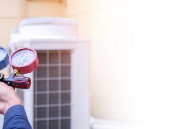 Close-up of a person using a manifold gauge set for a pressure reading on an air conditioning unit. The background shows the exterior of a building, emphasizing the importance of preventative maintenance plans for optimal performance.